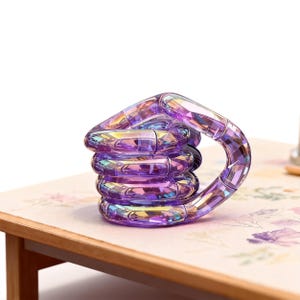 May include: A stack of five purple, faceted, interlocking rings forming a handle shape. The rings have a rainbow iridescence. The object is on a floral patterned surface, with a wooden table edge visible.