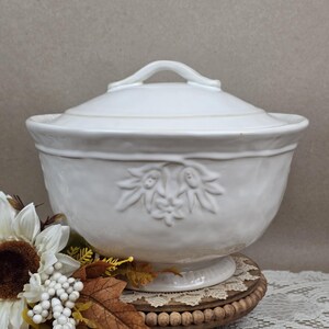May include: A white ceramic tureen with a matching lid, featuring a raised floral relief pattern on the front. The bowl has a rounded body, a flared rim, and a pedestal base. The lid includes a curved handle at the top for easy removal.