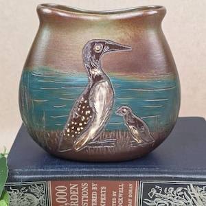 May include: A brown and teal ceramic vase featuring an embossed illustration of a large bird and a smaller bird standing in a grassy area near water. The vase has a curved, wide-mouthed opening and a smooth, matte finish on its exterior surface.