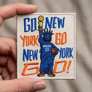 May include: A square embroidered patch on a cream background featuring a blue Statue of Liberty figure holding a gold trophy and a basketball. The text reads GO NEW YORK GO NEW YORK GO! in blue and orange thread, with NEW YORK 11 on the figure's chest.