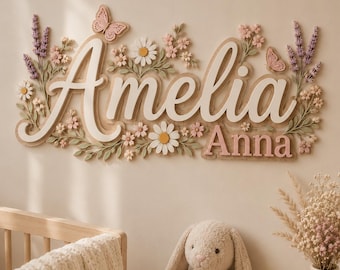 以下が含まれることがあります： AmeliaとAnnaの名前がレイヤー状の筆記体とブロック体で記された壁掛けサイン。白いデイジー、ピンクの花、ラベンダーの小枝、2匹の蝶など、立体的な花の装飾が施されています。