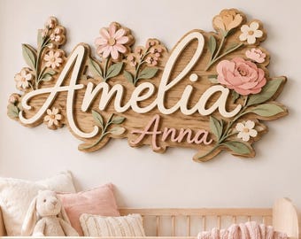 以下が含まれることがあります： 木製の壁掛け看板で、AmeliaとAnnaの名前が重なった筆記体で書かれています。ピンク、クリーム、セージグリーンの立体的な花の装飾が施され、柔らかい寝具を備えた木製のベビーベッドの上に設置されています。