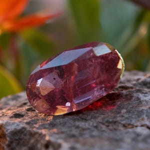 May include: A faceted oval gemstone with a deep magenta and purple hue. The stone features multiple geometric cut surfaces that reflect light, creating internal highlights and shadows. It is positioned on a rough, textured gray stone surface.