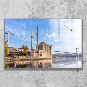 Op de afbeelding: Een glazen wandkunstprint met de Ortaköy-moskee in Istanbul, Turkije, met twee hoge minaretten en een centrale koepel. De Bosporusbrug overspant het water op de achtergrond, met meeuwen die in de helderblauwe lucht vliegen.