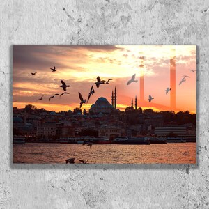 May include: A rectangular wall art print depicting a sunset over a city skyline with a large domed mosque and minarets. Numerous birds are flying across the orange and yellow sky above the water, where several boats are docked near the shore.