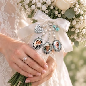 Pode incluir: Um pingente para buquê de noiva com três molduras ovais prateadas, um pingente em forma de coração com o texto 'you always in my heart' e contas de pérolas brancas. O pingente está preso a uma fita de cetim branca em um buquê de flores.