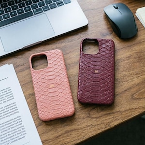 May include: Two phone cases with a snakeskin texture are displayed on a wooden surface. One case is a light pink, and the other is a deep burgundy. Each case has a cutout for the camera and a monogram. A laptop, mouse, and paper are in the background.