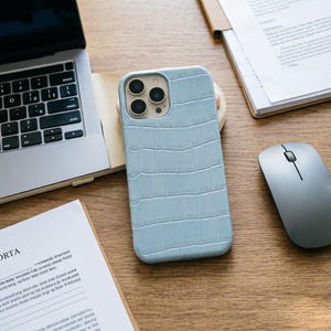 May include: A light blue phone case with a crocodile skin texture is displayed on a wooden desk. A laptop, a computer mouse, and documents are also on the desk. The phone case has a camera cutout and is next to a small wooden coaster.
