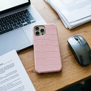 May include: A light pink phone case with a crocodile skin texture. The case is next to a laptop, a computer mouse, and some papers on a wooden desk. The phone case has three camera lenses.