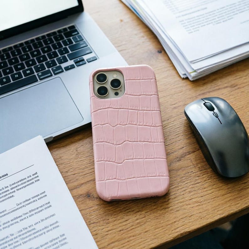 May include: A light pink phone case with a crocodile skin texture. The case is next to a laptop, a computer mouse, and some papers on a wooden desk. The phone case has three camera lenses.