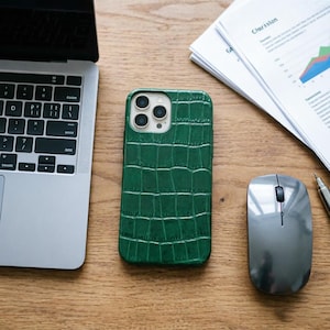 May include: A green textured phone case with a crocodile skin pattern. The phone case is on a wooden desk next to a laptop, a computer mouse, and a pen. The phone case has a camera cutout.