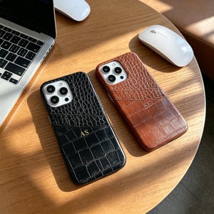 Pode incluir: Duas capas para smartphone com padrão de textura de pele de crocodilo, uma em preto com as iniciais AS gravadas em dourado e uma em castanho com as iniciais SE gravadas em dourado. Cada capa possui um bolso para cartões e abertura.