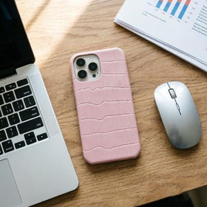 May include: A light pink phone case with a crocodile skin texture. The case is next to a silver computer mouse and a laptop on a wooden surface. The phone case has a camera cutout with three lenses.