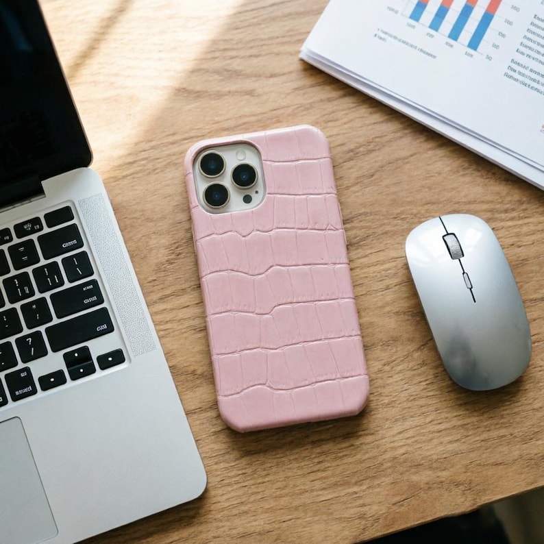 May include: A light pink phone case with a crocodile skin texture. The case is next to a silver computer mouse and a laptop on a wooden surface. The phone case has a camera cutout with three lenses.