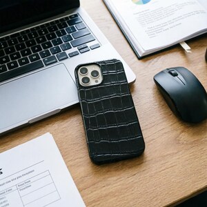 May include: A black textured phone case with a crocodile skin pattern rests on a wooden desk next to a laptop, a mouse, and a notebook. The phone case has a camera cutout and is designed to fit a smartphone.
