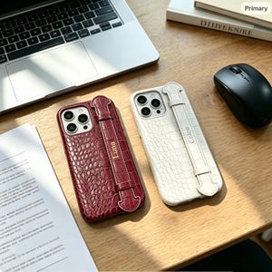 May include: Two smartphone cases with a crocodile-embossed texture, one in deep red and one in white. Each case features a vertical strap across the back with a gold-toned nameplate, displaying the names Luna on the red case and Chloe on the white case.