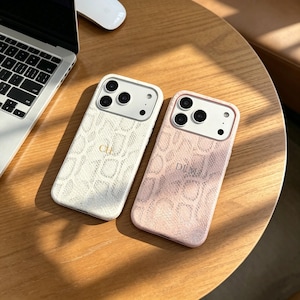 Pode incluir: Duas capas de smartphone com um padrão texturizado de pele de cobra. A capa da esquerda é branca com as letras douradas CH, e a da direita é rosa claro com as letras douradas DLM. Ambas têm um recorte branco para três lentes.