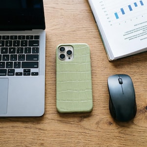 May include: A light green phone case with a crocodile skin texture rests on a wooden desk next to a laptop, a wireless mouse, and a book with charts. The phone case has three camera lenses.