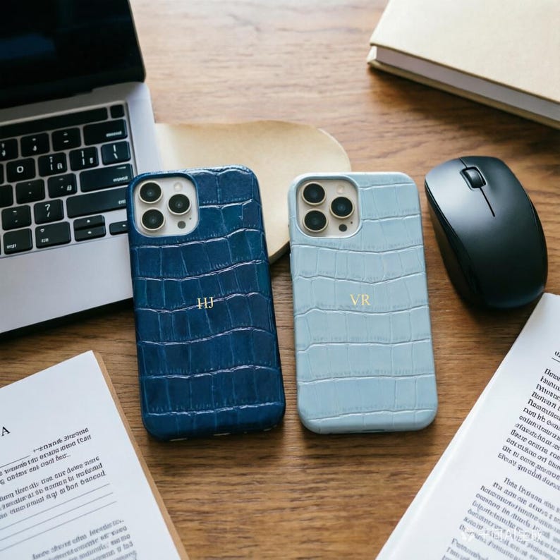 May include: Two leather phone cases with a crocodile texture, one navy blue and one light blue, are displayed on a wooden desk. Each case has gold initials. A laptop, mouse, and papers are also on the desk.