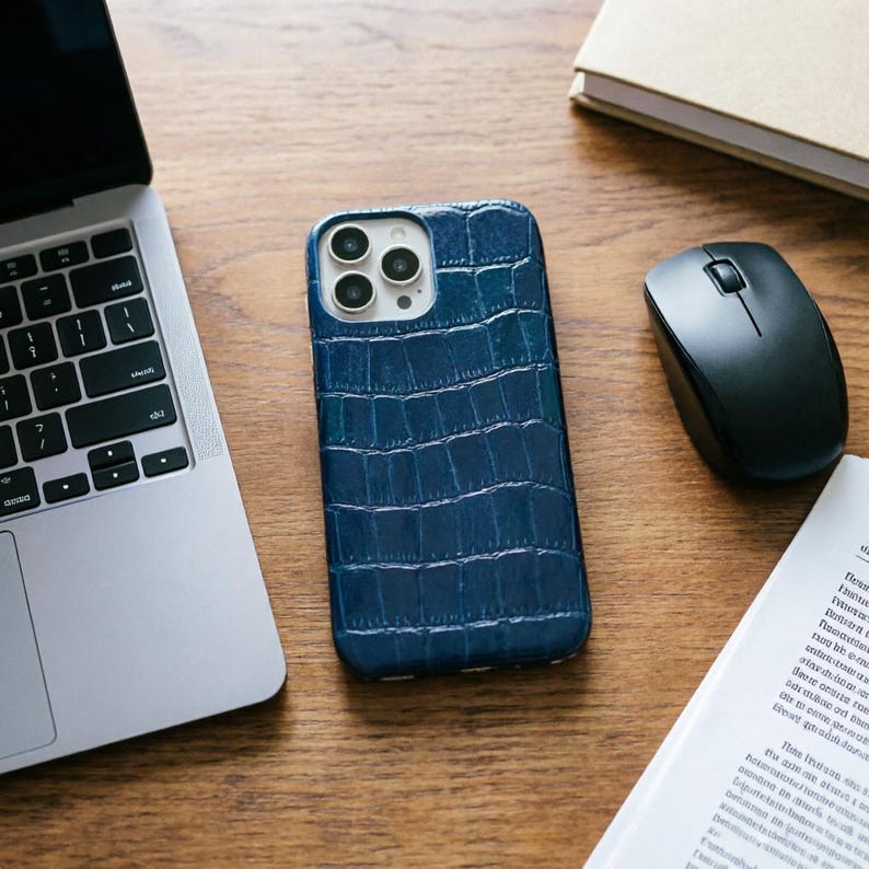 May include: A blue textured phone case with a crocodile skin pattern. The case is next to a laptop, a black computer mouse, and a book on a wooden surface.