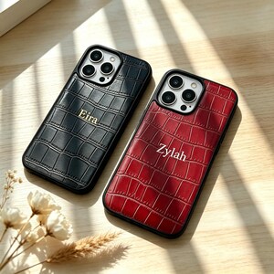 May include: Two smartphone cases with a textured crocodile skin pattern. One case is black with the name Eira printed in gold, and the other is deep red with the name Zylah printed in white. Both cases feature a triple camera cutout.
