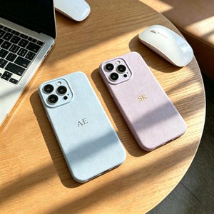 May include: Two smartphone cases with a textured finish, one in light blue with the gold embossed letters AE and one in light purple with the gold embossed letters SK. Both cases feature a square camera cutout with three circular lenses.