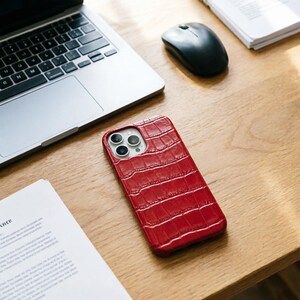 May include: A red, textured phone case with a crocodile skin pattern rests on a wooden desk. The case has a camera cutout and is next to a laptop, mouse, and documents. The case is designed to protect a smartphone.