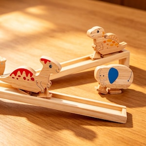 May include: Wooden toy set featuring a dinosaur and elephant rolling down wooden tracks. The dinosaur toys have red and orange accents. The elephant has a blue ear. The tracks are set on a wooden surface.