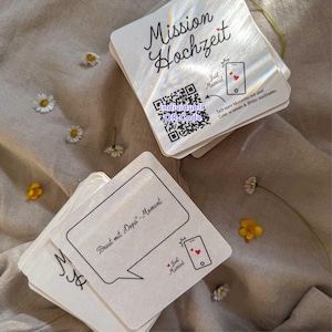 Könnte beinhalten: Ein Stapel quadratischer weißer Karten mit holografischem Finish. Die oberste Karte zeigt den Text Mission Hochzeit, einen QR-Code, ein Smartphone-Symbol mit Herzen und den Text Just Married. Eine weitere Karte zeigt eine Sprechblase mit Braut mit Oops-Moment.
