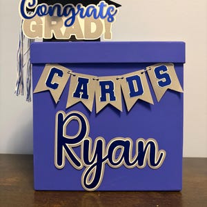 May include: A blue graduation card box with the name "Ryan" in white and blue lettering. The box has a banner that says "CARDS" and a graduation cap with the words "Congrats GRAD!" in blue and white.