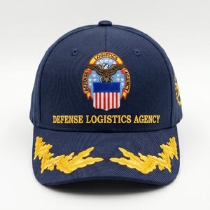 May include: A navy blue baseball cap featuring the embroidered Defense Logistics Agency emblem on the front. The emblem displays an eagle above a shield with red and white stripes. The text Defense Logistics Agency is stitched in yellow below.