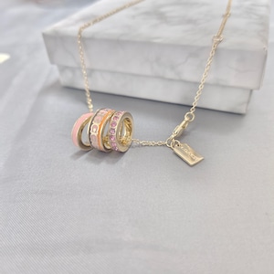 Puede incluir: Collar dorado con un colgante con tres anillos entrelazados. Los anillos son en tonos rosa y naranja, con un anillo adornado con piedras preciosas rosas. Una pequeña etiqueta rectangular con texto cuelga cerca del cierre.