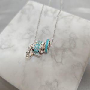 May include: A silver necklace featuring three decorative rings. One ring is encrusted with clear crystals, another has a turquoise and white patterned design, and the third is solid turquoise. The necklace hangs on a delicate silver chain.