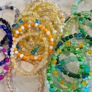 May include: A collection of elastic beaded bracelets featuring various colors including yellow, orange, green, blue, and pink. The bracelets are made with round beads, some translucent and some opaque, accented with small gold-toned metal spacers.