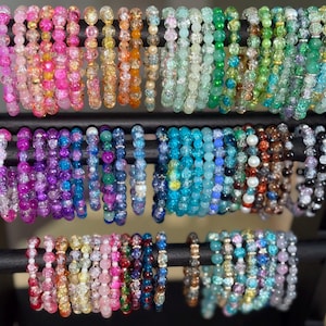 May include: A collection of numerous elastic bracelets featuring round, crackle-textured glass beads in a wide spectrum of colors including pink, yellow, orange, green, blue, purple, and clear, displayed on a black tiered jewelry stand.