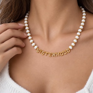 Puede incluir: Un collar de cuentas compuesto por esferas blancas lisas intercaladas con pequeños espaciadores metálicos dorados. En el centro, el collar presenta cuentas con letras doradas que forman la palabra SISTERHOOD en una fuente clara y sencilla.