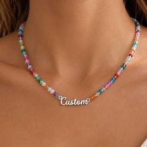 Puede incluir: Un collar que presenta una hilera de pequeñas cuentas translúcidas multicolores en tonos azul, verde, rosa, naranja y amarillo, centrado con una placa de metal plateado que muestra la palabra Custom en letra cursiva.