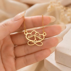 May include: A gold-toned necklace featuring a pendant with two interlocking heart outlines and an infinity symbol at the base. The names Aiden and Evelyn are engraved in a cursive script within each heart. The pendant hangs from a delicate gold chain.