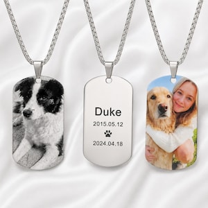 Pode incluir: Três pingentes de placa de identificação prateados em correntes de elos quadrados correspondentes. O pingente esquerdo apresenta uma foto em preto e branco de um cão. O central está gravado com o nome Duke, as datas 2015.05.12 e 2024.04.18 e uma pata. O pingente direito mostra uma foto colorida de uma pessoa abraçando um cão dourado.