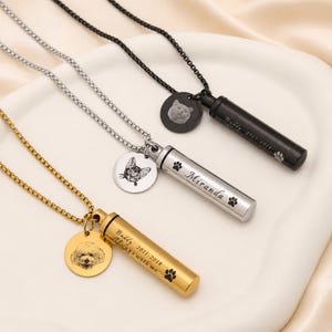 Pode incluir: Três colares com pingentes cilíndricos em tons de ouro, prata e preto. Cada um apresenta um berloque circular gravado com o retrato de um animal e um pingente de tubo gravado com nomes, datas e pegadas em cores correspondentes.