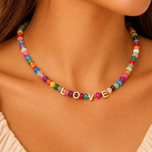 May include: A beaded necklace featuring a sequence of small, multicolored, translucent circular beads. The necklace includes four gold-toned metal letter charms positioned in the center that spell out the word LOVE in capital letters.