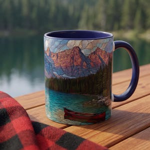 May include: A ceramic mug with a dark blue interior and handle, featuring a stained-glass style wrap-around print of a red canoe floating on a turquoise lake, surrounded by a dense evergreen forest and tall, jagged mountain peaks under a cloudy sky.