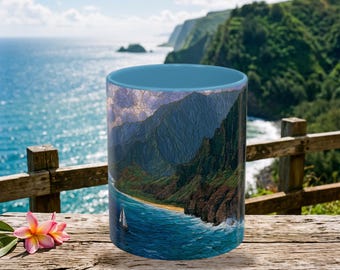 May include: A ceramic mug with a light blue interior and rim, featuring a wrap-around graphic print of a coastal landscape with steep green cliffs, a sandy beach, blue ocean water, and a small white sailboat under a cloudy sky.
