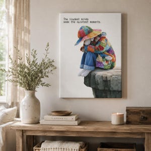 Op de afbeelding: Een rechthoekige canvas print met een figuur in een kleurrijke patchwork hoed en jas, zittend op een rotsachtige richel. De tekst "The loudest minds seek the quietest moments." staat in het zwart gedrukt in de linkerbovenhoek.