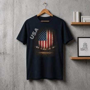 May include: A dark navy blue short-sleeved t-shirt featuring a graphic print of the United States flag centred on the chest. The word USA is printed in white capital letters vertically along the left side of the flag graphic, which has a dark, textured background.