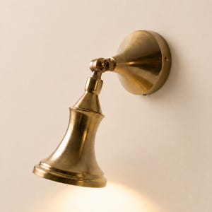 Puede incluir: Un aplique de pared con acabado en latón pulido, pantalla cónica y base circular. La lámpara incluye una articulación giratoria ajustable que conecta la pantalla con la placa de montaje, permitiendo orientar la luz de forma direccional.