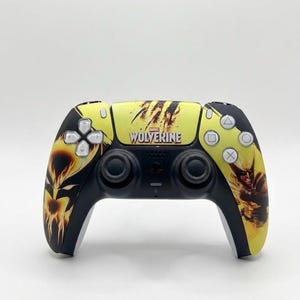 May include: A black and yellow video game controller featuring a Marvel Wolverine graphic. The center displays the Wolverine logo with claw marks, while the right side shows a character illustration. It includes standard buttons, joysticks, and a D-pad.