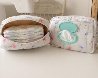 May include: Two quilted fabric pouches featuring a delicate floral print in shades of pink, blue, and green. One pouch is open, revealing a stack of folded white diapers inside. The other pouch is closed and has a light blue plastic dispenser lid.