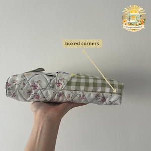 May include: A quilted pouch featuring a white fabric with a pink and purple floral pattern on one side and a green and white gingham check pattern on the other. The pouch has a rectangular shape with boxed corners and a zipper closure at the top.