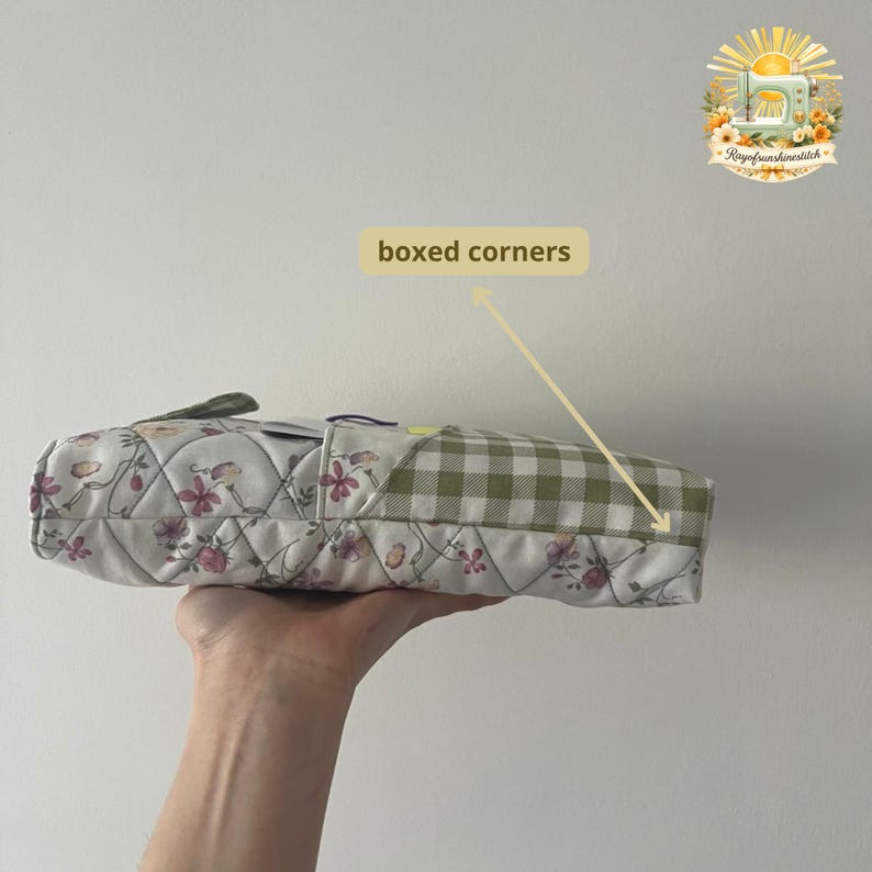 May include: A quilted pouch featuring a white fabric with a pink and purple floral pattern on one side and a green and white gingham check pattern on the other. The pouch has a rectangular shape with boxed corners and a zipper closure at the top.
